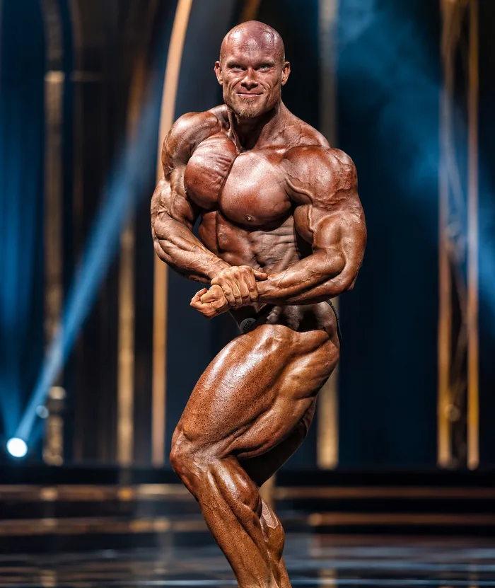 A muscular bodybuilder poses under stage lighting.